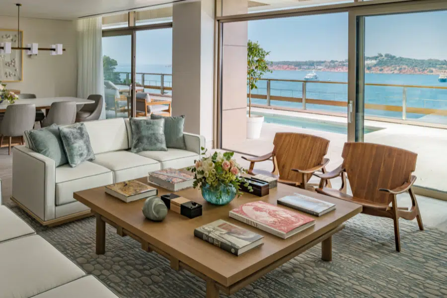 Modern living room with neutral-toned sofas, wooden chairs, and a coffee table with books and flowers, overlooking a balcony with a pool and ocean view through large glass doors.