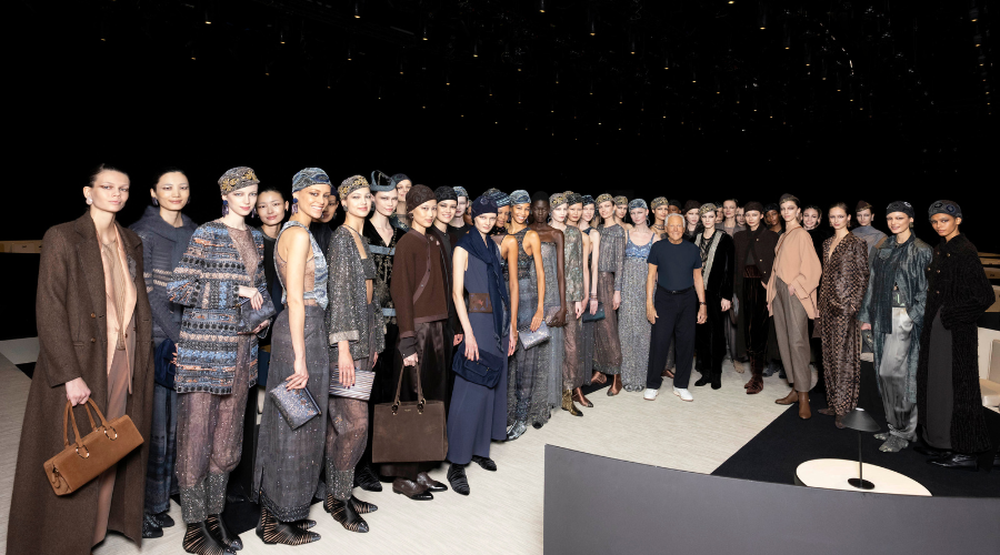 A group of fashion models wearing coordinated, elegant outfits and headbands pose together on a runway with two people standing among them, under soft lighting in a spacious, dark venue.
