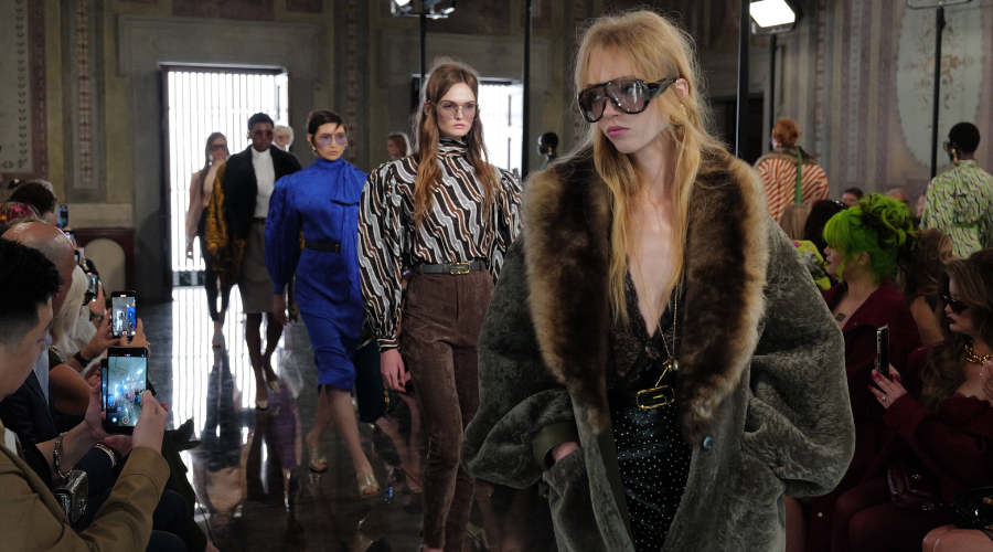Models walk down a fashion runway wearing stylish outfits, including a fur coat and striped blouse, as audience members take photos with their phones in an elegant, ornately decorated room.