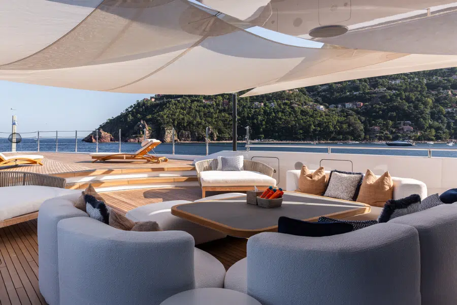A luxurious yacht deck with modern white sofas, a wooden table, sun loungers, and shaded canopies overlooks calm blue water and a lush, green hillside.