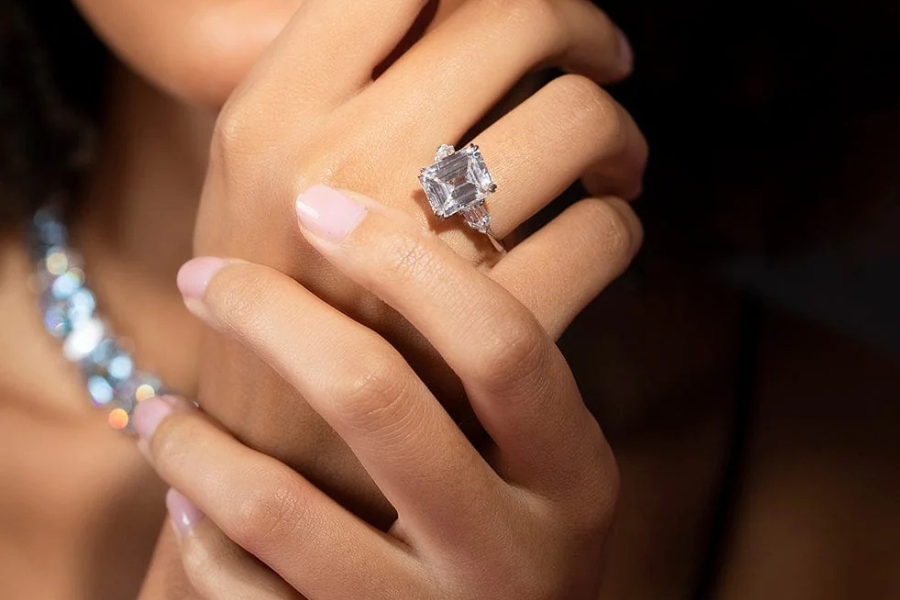 A close-up of a womans hand with light pink nail polish, displaying a large, square-cut diamond ring on her finger. A sparkly necklace is visible in the blurred background.