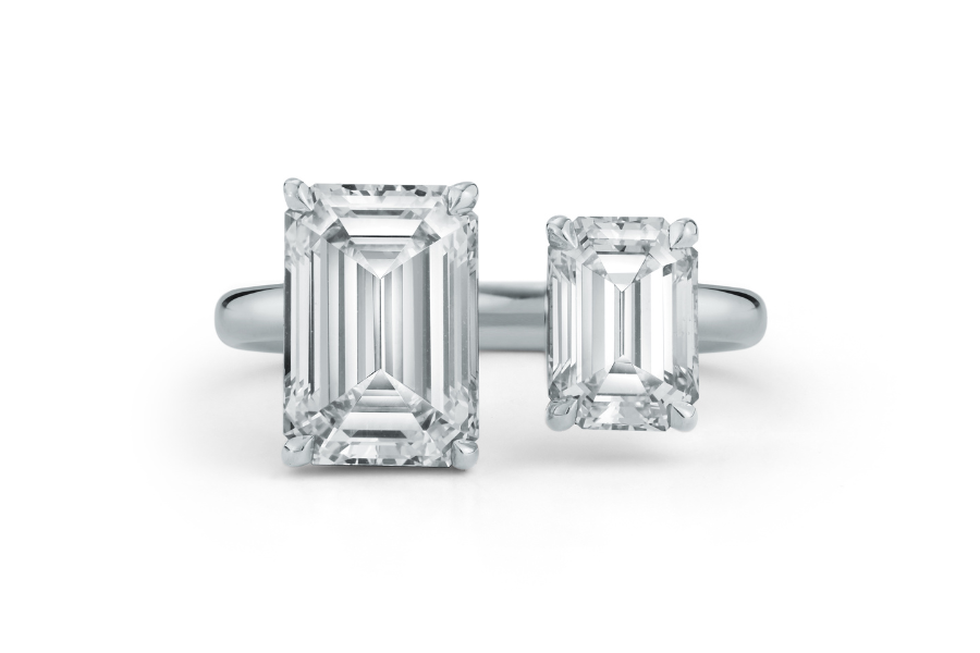 A silver ring featuring two emerald-cut white diamonds of different sizes side by side, set on a simple band against a white background.