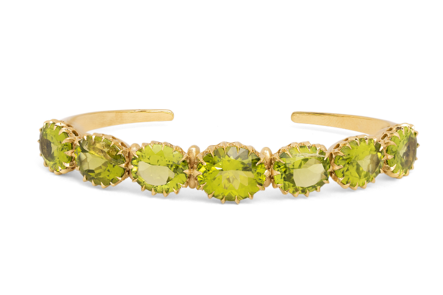 A gold cuff bracelet featuring a row of seven oval-cut green gemstones, each set in a prong setting, displayed against a white background.