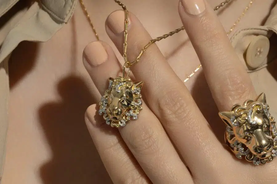 A close-up of a hand with nude nail polish wearing a gold lion head ring and holding a matching lion head pendant necklace with sparkling stone accents. The person is dressed in a beige shirt.