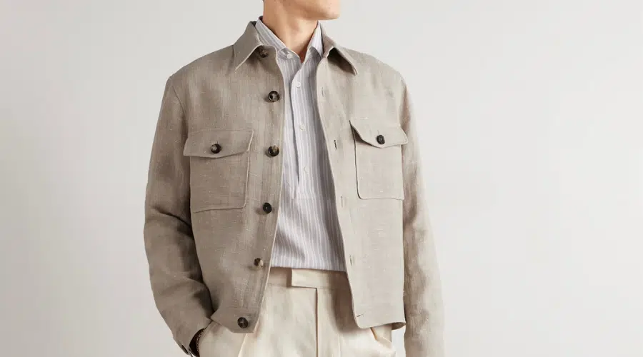 A person wearing a light beige button-up jacket with flap pockets over a light striped shirt and cream trousers, standing with one hand in their pocket against a plain background.