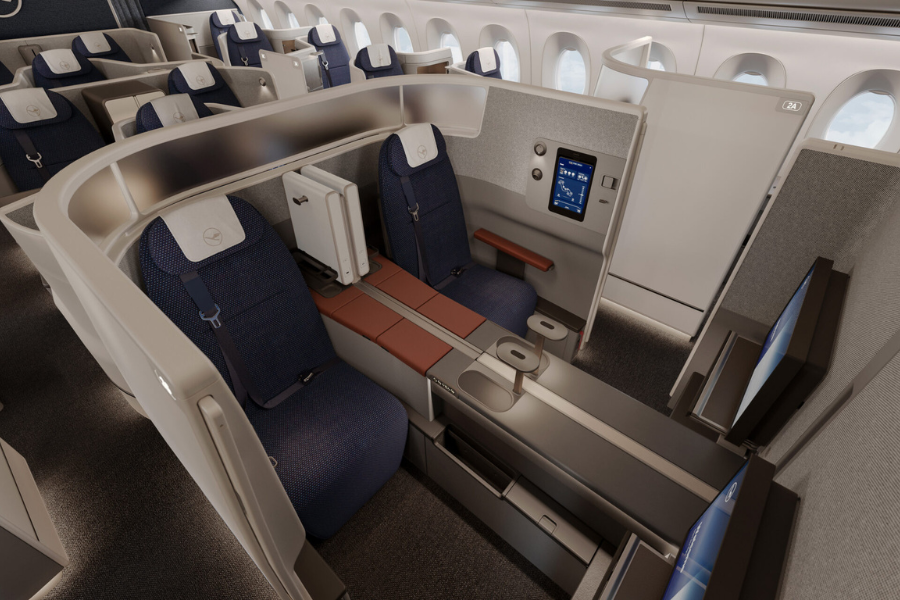 A modern airline business class cabin featuring spacious seats with privacy dividers, large entertainment screens, and ample legroom. Seats are upholstered in dark blue fabric with tan and brown accents.