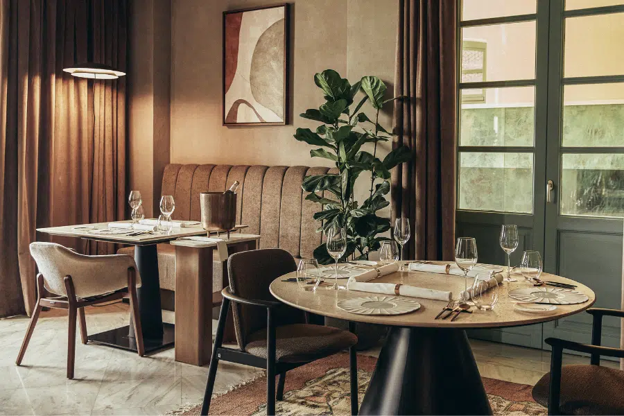 A cozy, stylish restaurant interior with set tables, cushioned chairs, wine glasses, a potted plant, abstract wall art, and soft natural light coming through large windows with curtains.