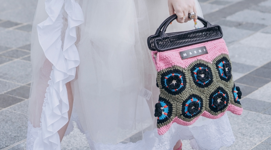 A person in a white ruffled dress holds a colorful Marni crochet handbag with pink, green, black, blue, and white circular patterns while standing on a tiled sidewalk.