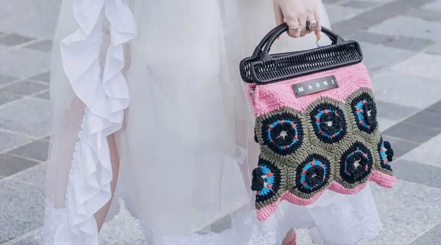 A person in a white ruffled dress holds a colorful Marni crochet handbag with pink, green, black, blue, and white circular patterns while standing on a tiled sidewalk.