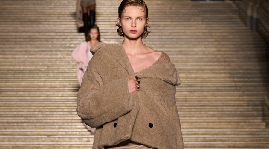 A model walks down stone steps in an oversized beige coat with large buttons, styled off the shoulders, during a fashion show; another model is visible in the background.