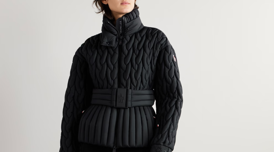 A person wearing a black, quilted, high-collar puffer jacket with a wide belted waist, standing against a plain light background.