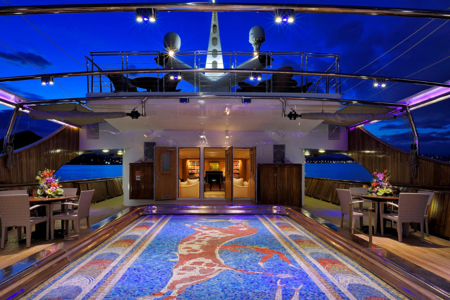 A luxurious yacht deck at dusk features elegant dining tables, floral centerpieces, wooden paneling, and a colorful tiled floor with a fish design, all illuminated by soft blue and purple lighting.