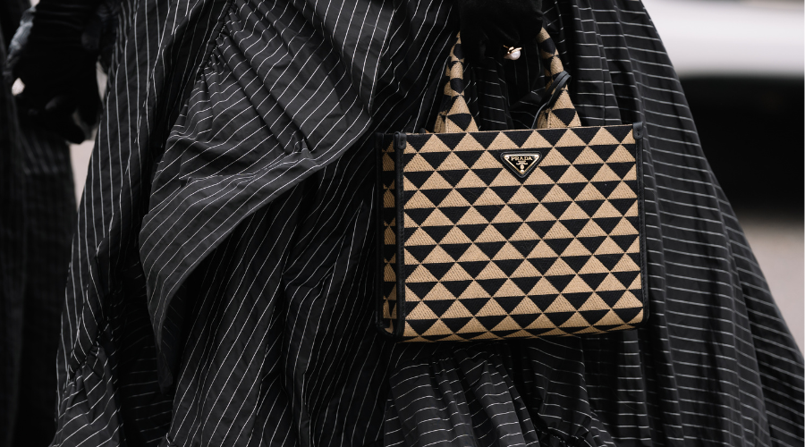 A person in a black pinstripe outfit holds a tan and black Prada handbag with a bold geometric triangle pattern, shown in close-up.