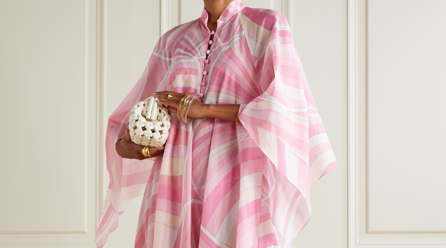 A person wears a flowing pink and white striped kaftan-style dress, accessorized with gold jewelry and holding a small white woven handbag, standing in front of a white paneled wall.