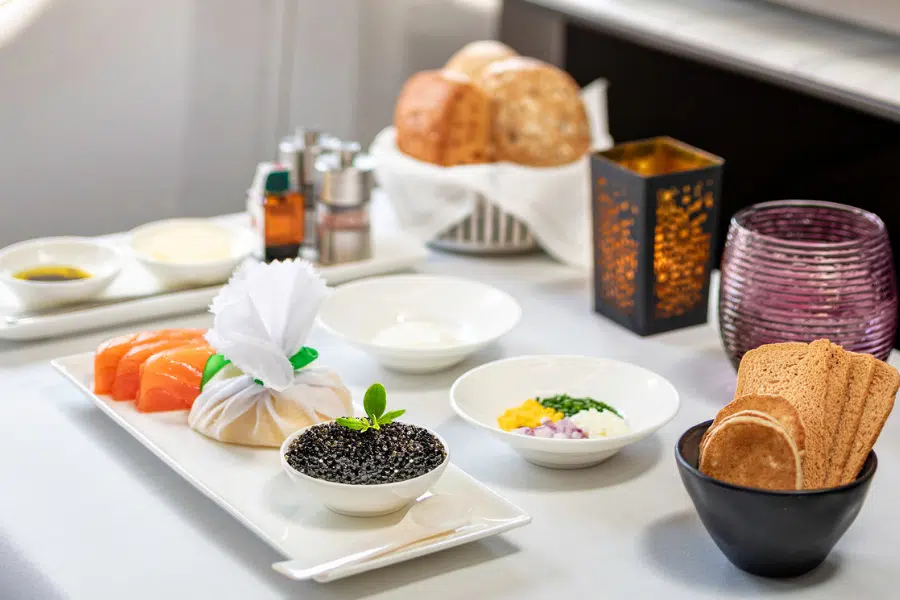 A table set with plates of smoked salmon, black caviar, crackers, assorted breads, butter, olive oil, and small bowls of diced onion, egg yolk, and egg white, with candles and condiments in the background.