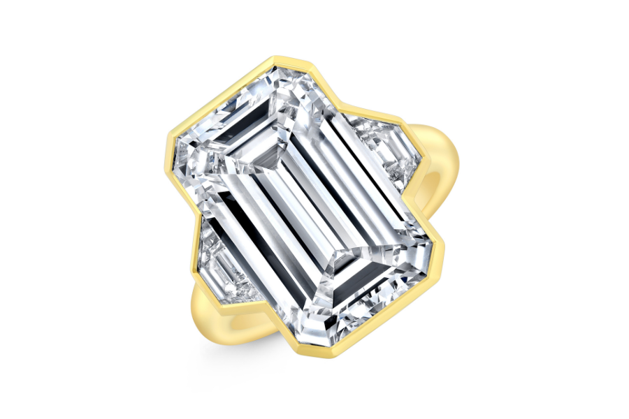 A gold ring featuring a large, uniquely-shaped emerald-cut diamond with multiple facets, set in a modern geometric design against a white background.