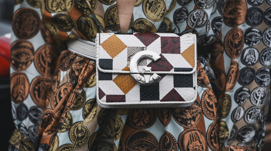 A close-up of a geometric-patterned handbag with a silver G clasp, worn over a dress featuring ornate circular designs in earthy tones.