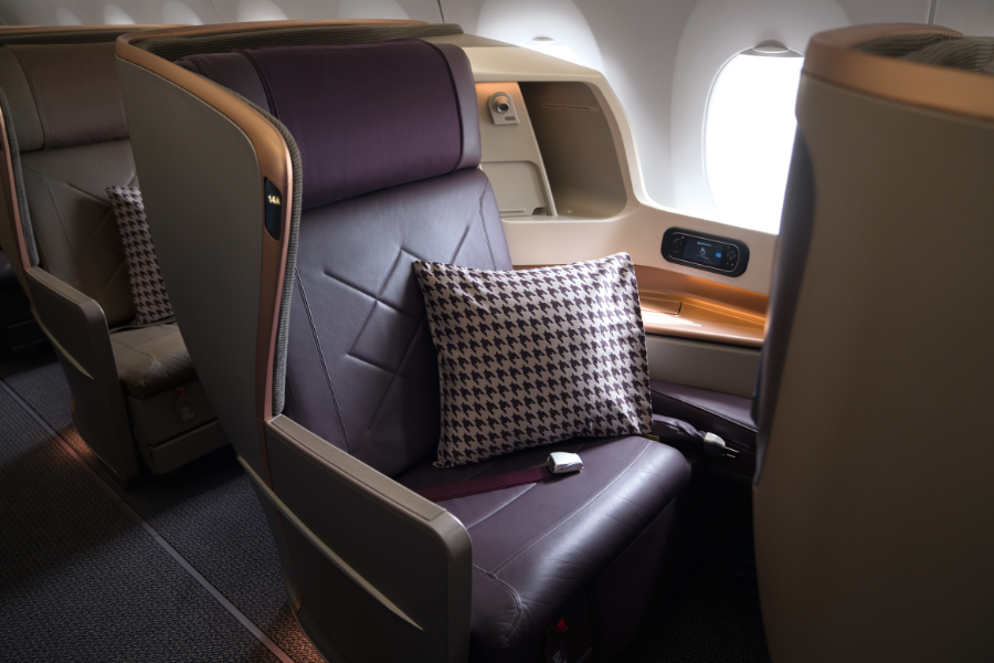 A luxurious airplane first-class seat with dark leather upholstery, a houndstooth-patterned pillow, seat controls, and a window nearby, showcasing a spacious and comfortable travel environment.