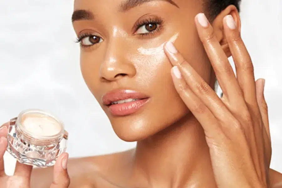 A woman applies a creamy skincare product to her cheek with her fingers, holding a small jar in her other hand. Her skin looks smooth and radiant.
