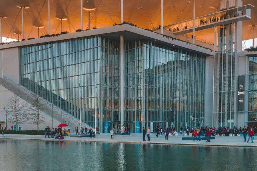 A modern glass building with tall windows and an overhanging roof, reflected in a large pool of water, with many people walking and gathering outside.