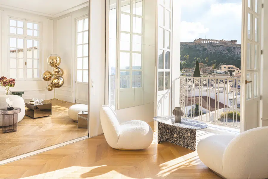 A bright, modern living room with curved white chairs, gold floor lamps, and a small marble table opens onto a balcony with a view of the Acropolis in Athens under a sunny sky.