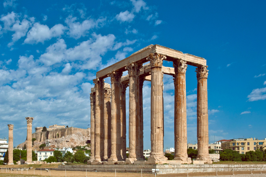 Ancient marble columns of the Temple of Olympian Zeus in Athens, Greece, stand under a blue sky with scattered clouds; the Acropolis is visible in the background.