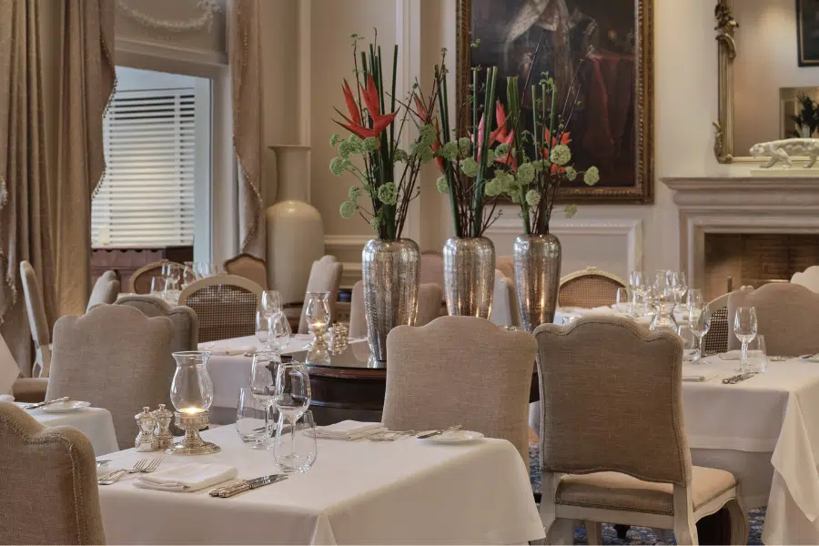 Elegant restaurant dining room with beige chairs, white tablecloths, and tables set with glassware and silverware. Three tall vases with red and green flowers are on a central table, and artwork hangs on the wall in the background.