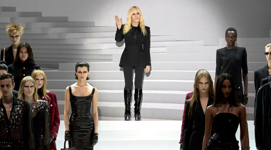 A blonde woman in black stands on white steps, waving, surrounded by fashion models in black outfits on a runway with a minimalist, monochrome background.