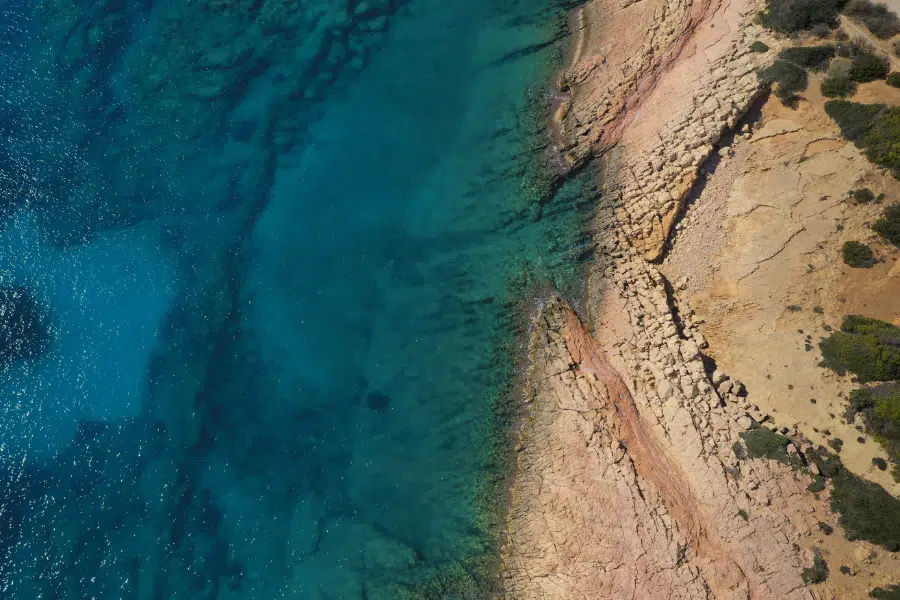 Aerial view of a rocky coastline with clear turquoise water on the left and rugged, sunlit brown cliffs and sparse vegetation on the right. The water gradually deepens away from the shore.