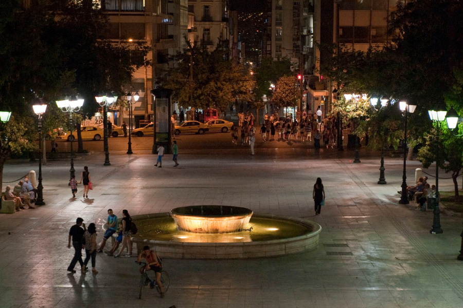 A city square at night with a lit fountain in the center, people walking and sitting on benches, streetlights illuminating the area, and buildings with lit windows in the background.