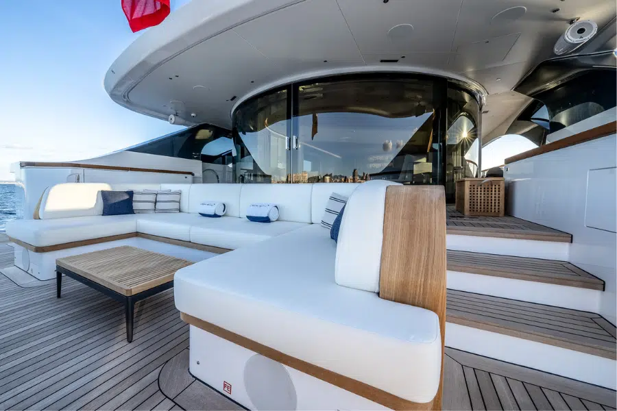 Modern yacht deck with white cushioned seating, wooden table, and steps leading to a glass-walled interior. Blue and white accent pillows decorate the seating area. Ocean and sky are visible in the background.
