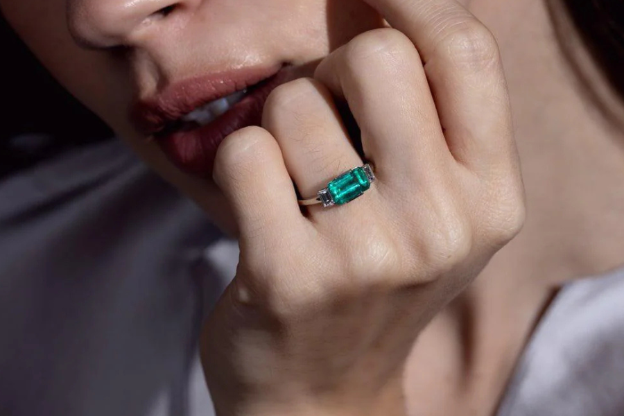 A close-up of a person’s hand near their lips, showing a silver ring set with a large rectangular green gemstone, possibly an emerald, on their finger. The lighting highlights the gemstone and the persons soft features.