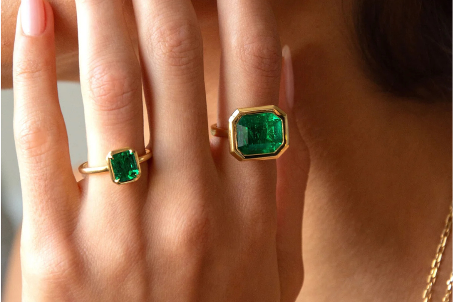 A close-up of a persons hand wearing two gold rings, each set with a large, rectangular green gemstone, possibly emeralds. The persons nails are manicured with pale polish, and their skin tone is medium.