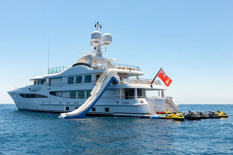 A large white luxury yacht anchored in calm blue water, featuring a slide leading from the upper deck into the sea. Several jet skis are lined up at the rear, and a red flag is flying from the yacht’s stern.