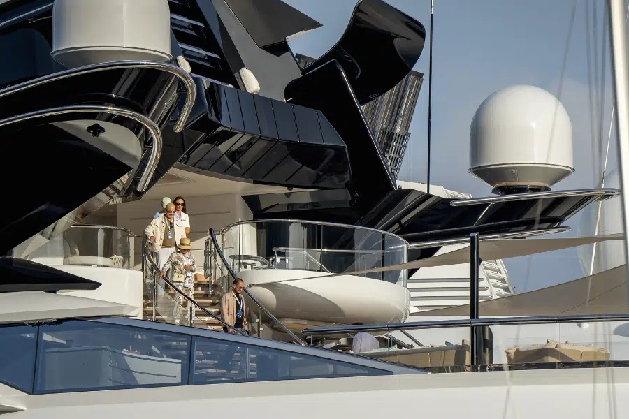 A group of people dressed in light-colored clothing stand on the deck of a sleek, modern yacht with glossy black and white surfaces and futuristic design elements, set against a blue sky.