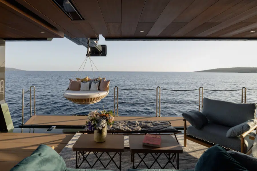 A luxurious yacht deck with a hanging round lounge chair, cushioned seating, a coffee table with flowers, and a view of the calm sea and distant hills under a clear sky.