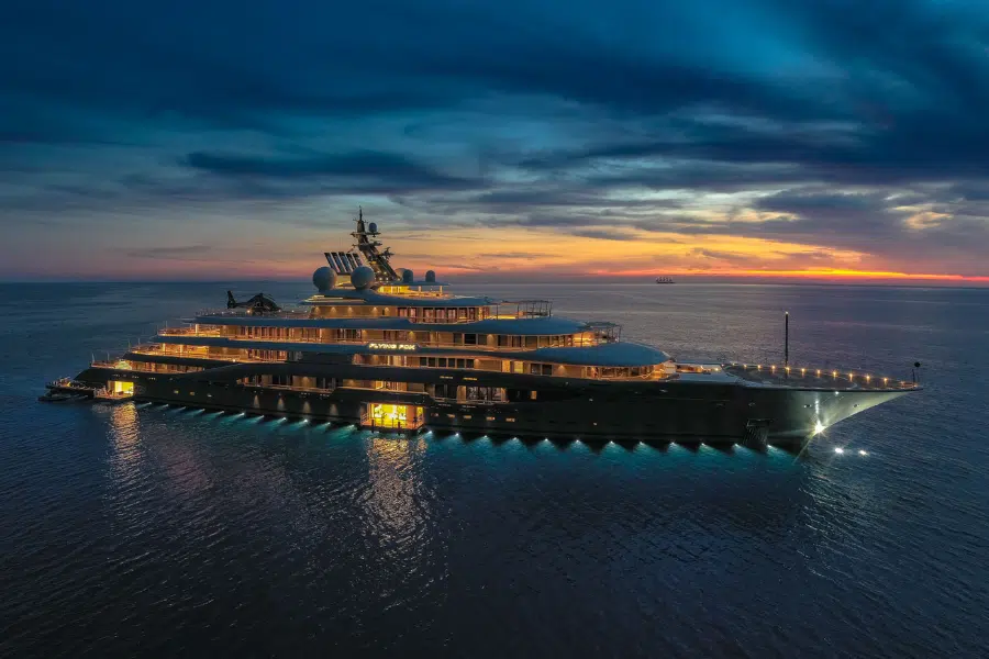 A large, brightly lit luxury yacht is anchored on calm water at sunset, with a dramatic sky and colorful clouds in the background.