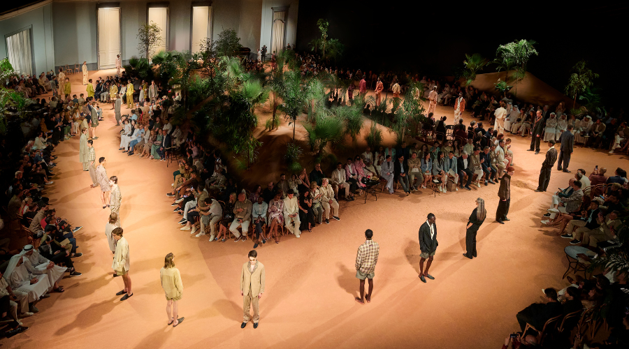 Models walk and stand on a circular runway surrounded by seated spectators, with tropical plants in the center, in a spacious, elegant indoor venue during a fashion show.