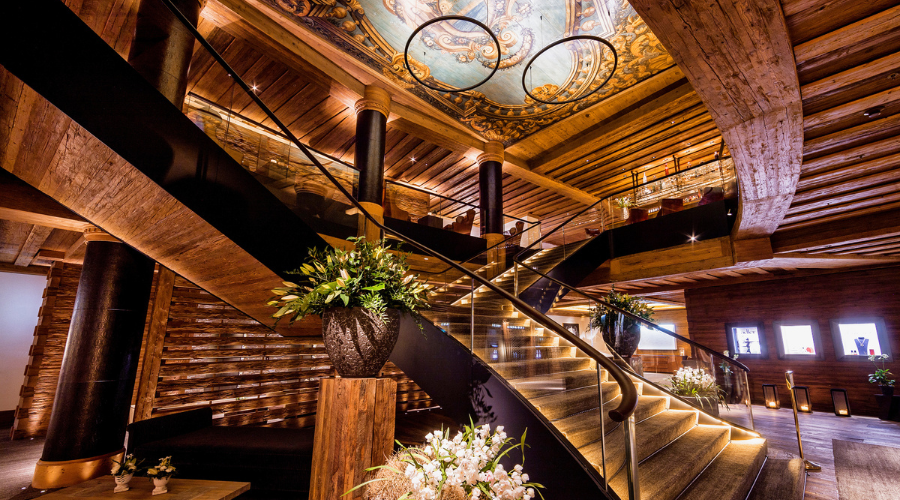 Elegant wooden interior with a grand curved staircase, glass railings, and ornate ceiling art. Large floral arrangements decorate the space, which is warmly lit with spotlights and accent lights along the steps.
