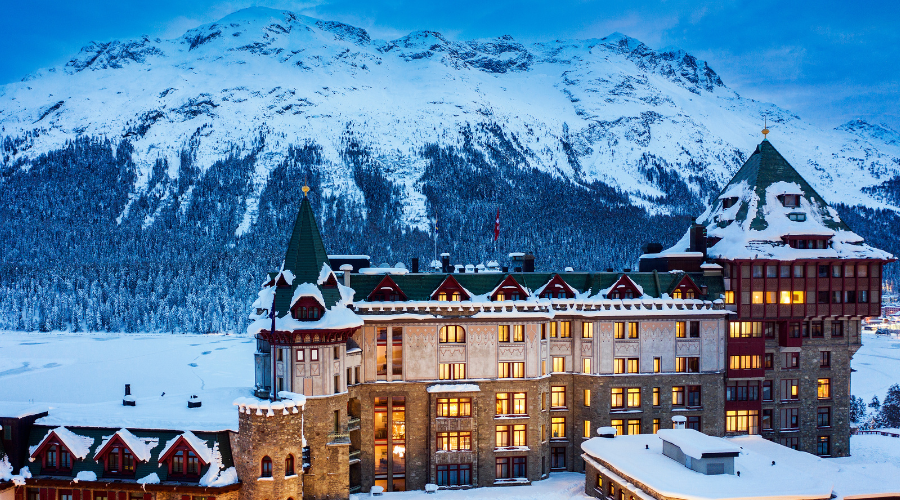 A grand, snow-covered hotel with green-roofed towers stands in front of a snowy mountain range and dense pine forest at dusk, with warm yellow lights glowing from the windows.