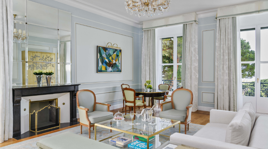 Elegant living room with pastel walls, a black fireplace, armchairs, a glass coffee table, books, and a sofa. Large windows with white curtains let in natural light. Modern art hangs above the fireplace.