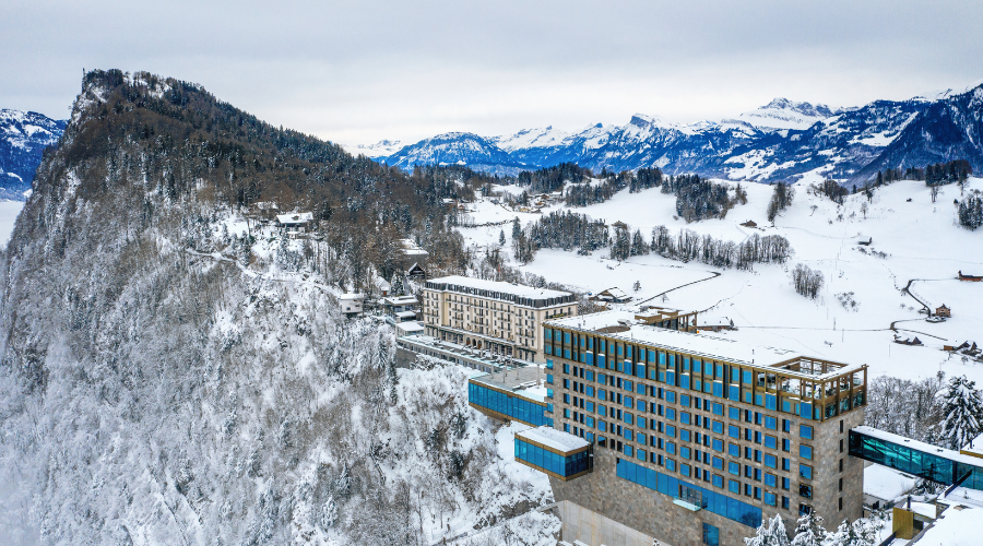 A modern hotel with glass windows stands on a snowy mountain slope, overlooking a winter landscape with distant mountains, trees, and scattered buildings under a cloudy sky.