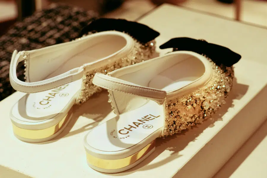 A pair of elegant Chanel slingback shoes with gold glitter, black bows on the back, and gold-trimmed heels, displayed on a white platform.