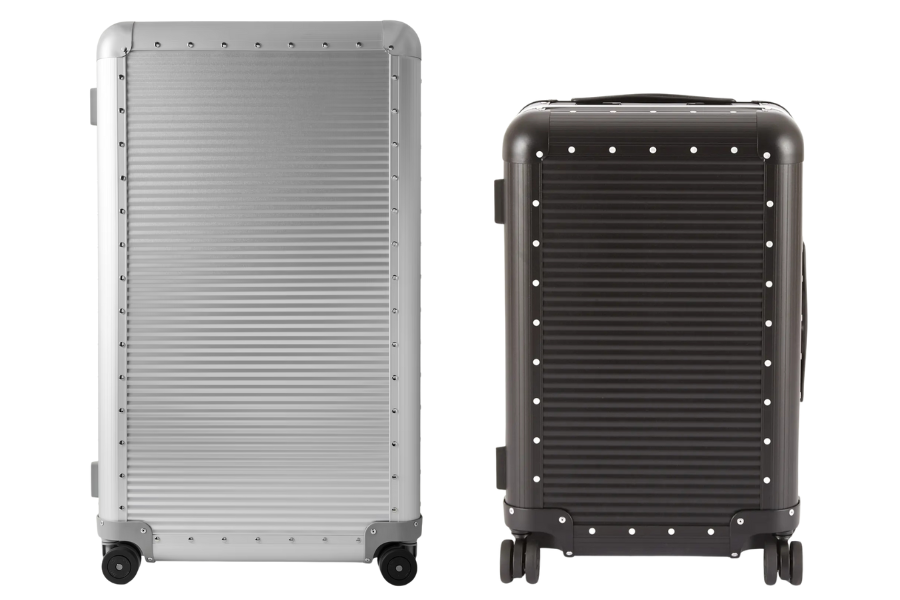 Two hard-shell suitcases stand side by side; the one on the left is silver with a ribbed texture, and the one on the right is black with a similar ribbed design. Both have four wheels and rivet accents around the edges.