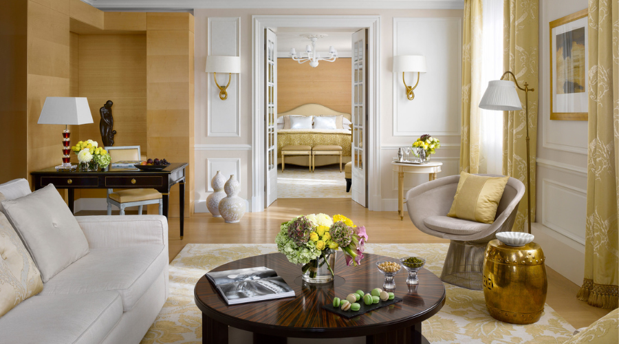 A bright, elegant living room with cream and gold tones features a sofa, round coffee table with flowers, armchair, and desk. Double doors open to a bedroom with a bed and chandelier in the background.
