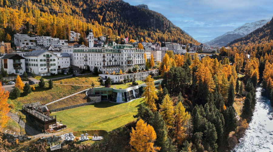 A grand hotel complex sits on a hillside surrounded by autumn-colored trees, next to a river, with mountains in the background under a partly cloudy sky. The scene is vibrant and picturesque.