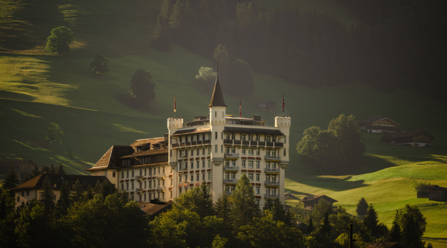 A grand, castle-like building with towers and flags stands among lush, green hills and trees, bathed in warm sunlight with forested mountains in the background.