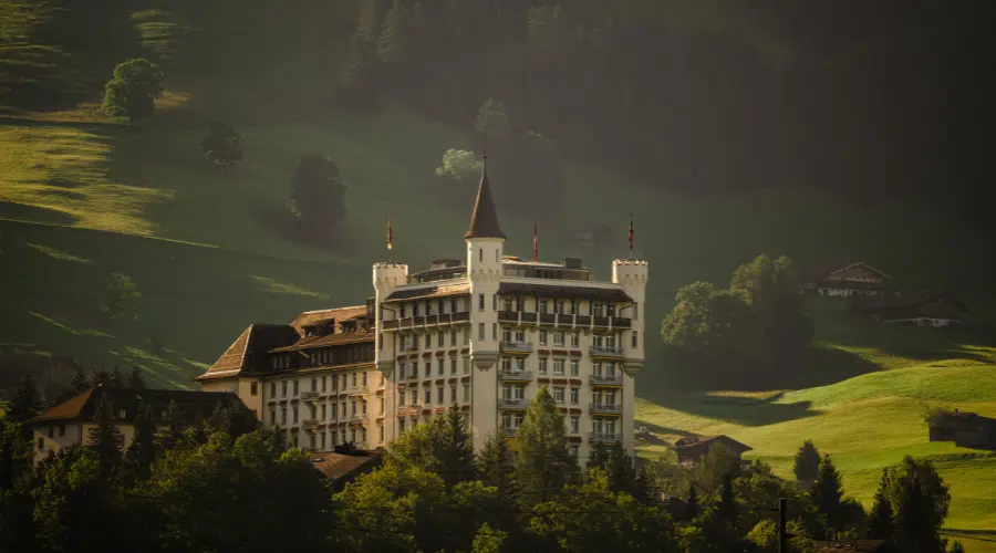 A grand, castle-like building with towers and flags stands among lush, green hills and trees, bathed in warm sunlight with forested mountains in the background.
