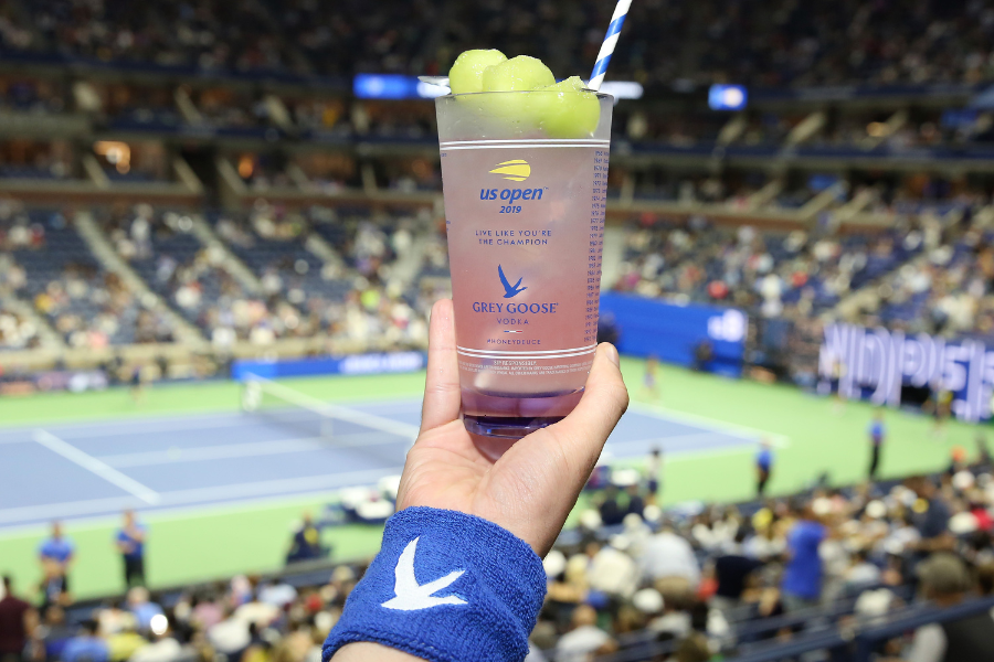 A hand wearing a blue wristband holds up a Grey Goose cocktail garnished with melon balls and a striped straw, with a tennis court and a crowd at the US Open in the background.