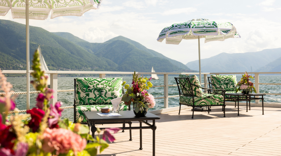 Outdoor lounge area with patterned green and white chairs and umbrellas, flower arrangements on tables, and a scenic view of a lake with sailboats and mountains in the background.